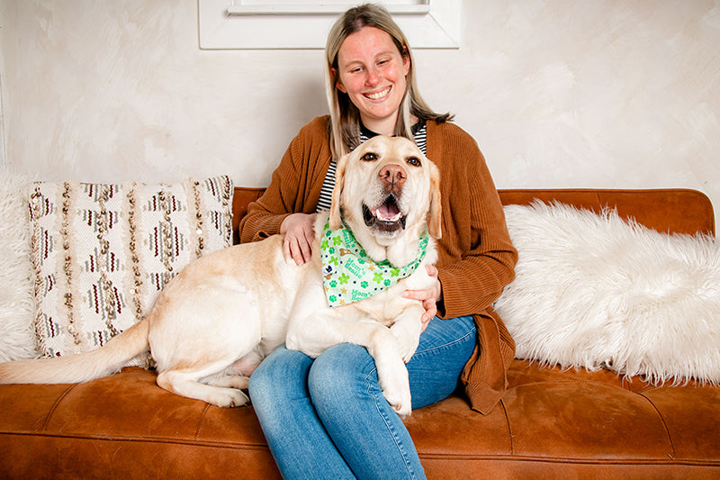 Woman sitting on a brown couch with a large white dog wearing a green mom's bestie bandana.