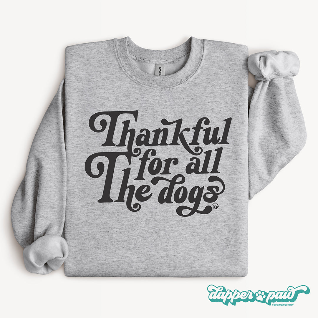 A folded sport gray sweatshirt with the phrase 'Thankful for all the dogs' printed in a large black decorative font on the front.