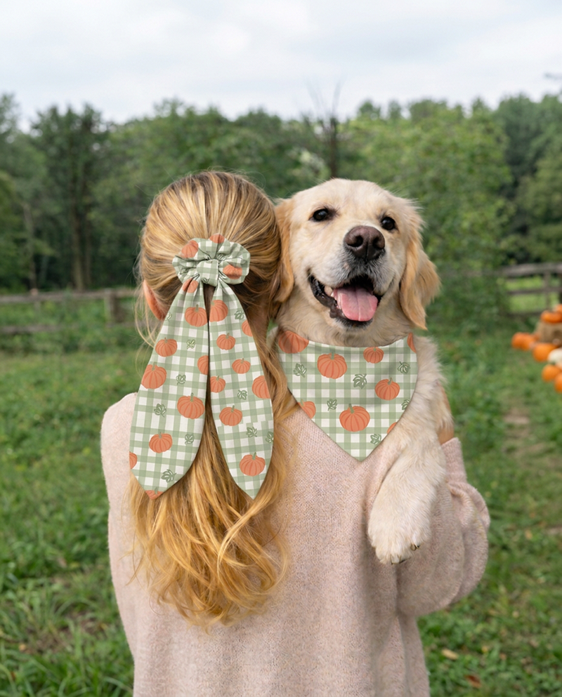 A woman and a golden retriever dog wearing matching green and orange pumpkin-print pet accessories, photographed outdoors in a grassy field with trees in the background.