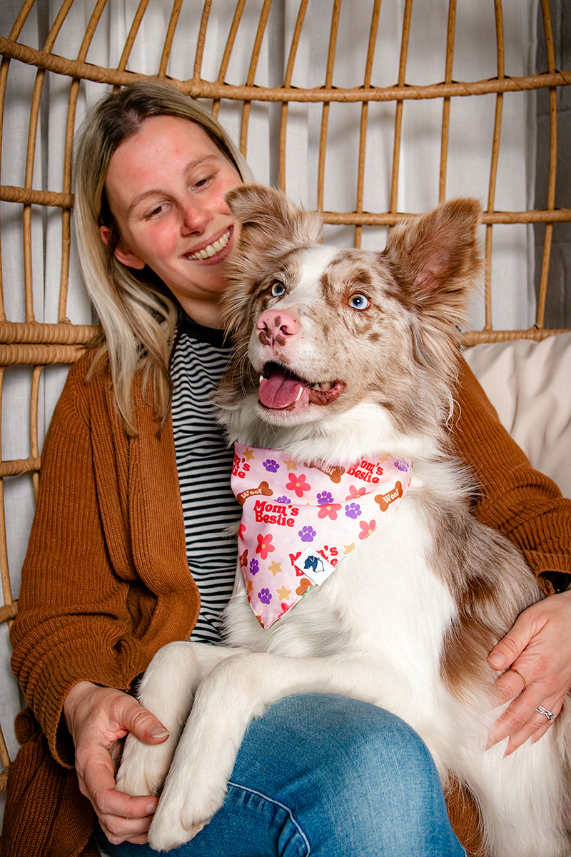 Woman holding a dog with a colorful bandana that says moms bestie in front of a wicker chair.